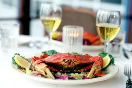 A Dungeness crab sits on a plate in a restaurant with two glasses of white wine in the background.